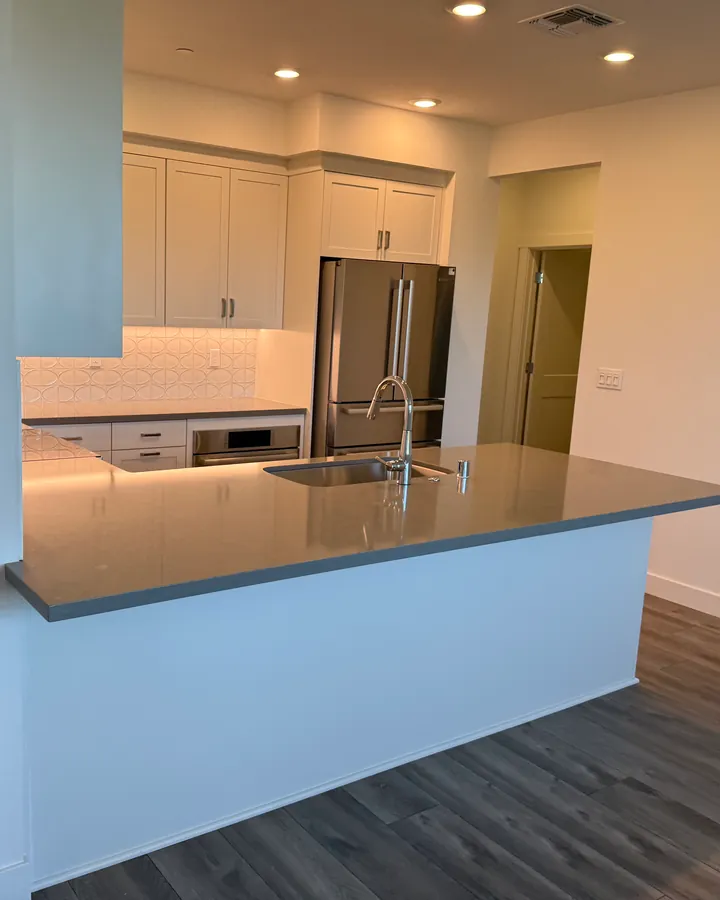 An empty apartment kitchen with white cabinets, a stainless-steel fridge and a long grey counter with the sink