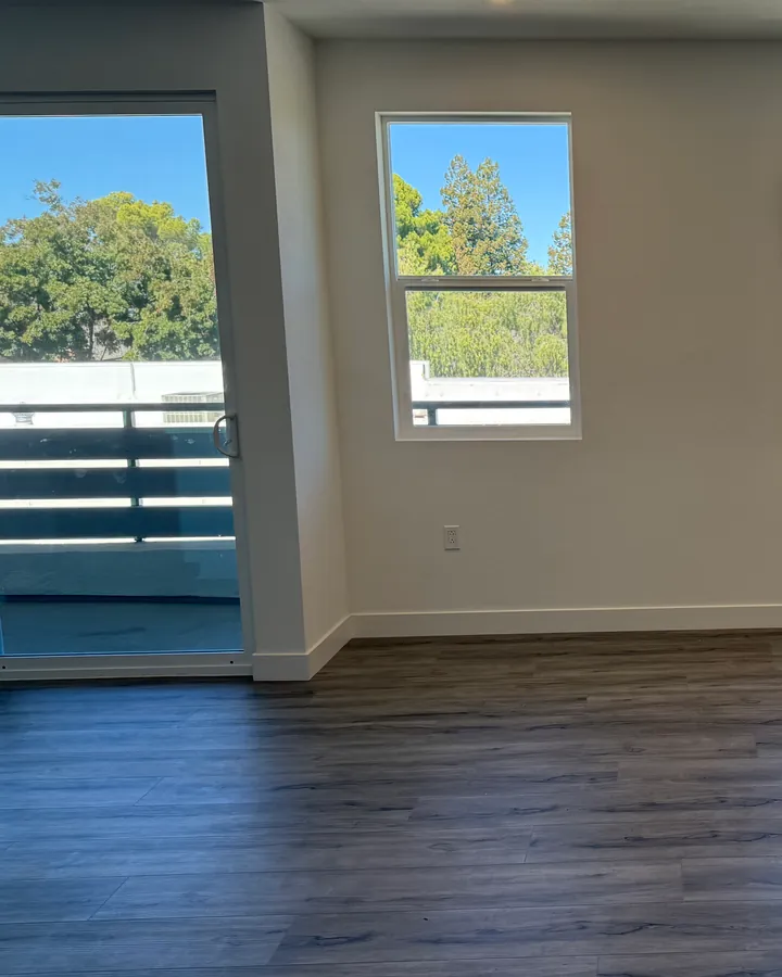An empty apartment living room with a sliding glass door to the balcony and a window looking out on trees